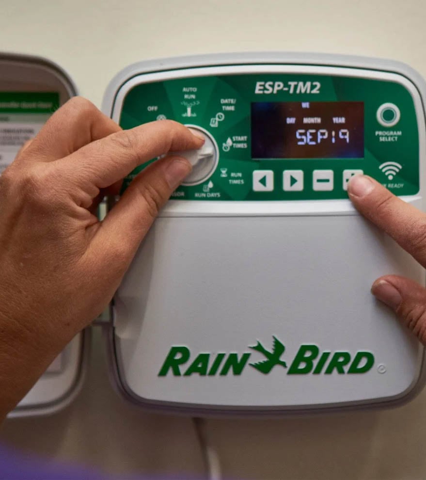 Hands adjust a Rain Bird ESP-TM2 irrigation controller with a dial, buttons, and an LCD screen.