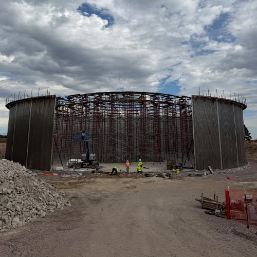 A large steel and concrete circular structure under construction. Four workers stand near the base wearing safety gear, and the walls rise above them, approximately 30-35 feet in height.