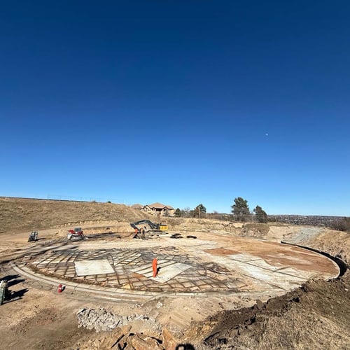 Worker stands in a large circular foundation of a structure, approximately 100 feet in diameter. The area is surrounded by recently excavated dirt under blue skies.