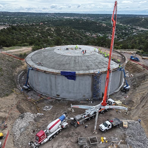 Aerial photo shows the finished side of a circular, domed, concrete water storage tank. A concrete truck and a large mechanism to pump cement to the roof of the structure are also visible.