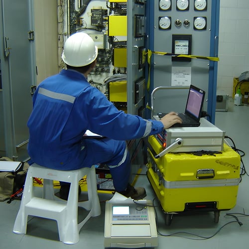 A person in a hardhat and work uniform sits in front of large machinery that contains circuitboards inside. They are using a laptop placed upon a yellow storage container to adjust equipment.