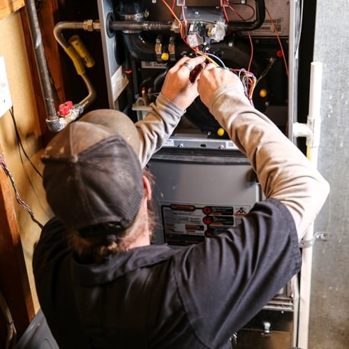 A man wearing a hat in a black shirt is fixing wires in a furnace. The panel of the furnace has been removed showing wires and pipes.