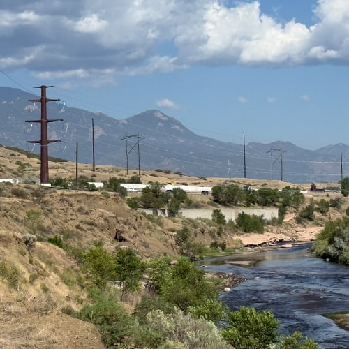 Large power lines and a brown metal transmission power pole is visible in the distance next to blue waters of Fountain Creek.
