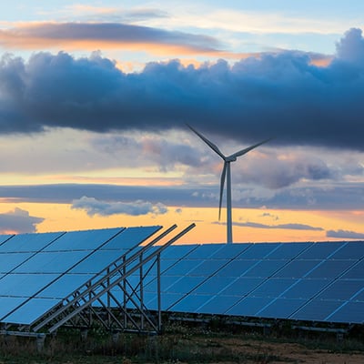 Solar panels in the foreground with a wind turbine visible in the distance at dusk.