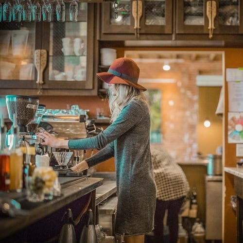 A person standing behind the bar at a coffee shop presses a button to grind coffee beans.