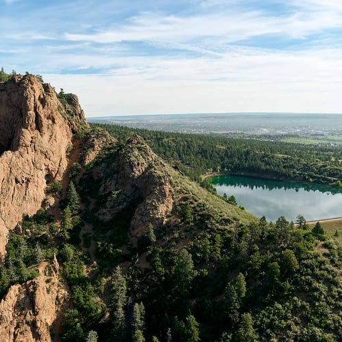 A rocky, tree covered ridge next to reservoir. The Colorado Springs skyline is faintly visible in the distance.