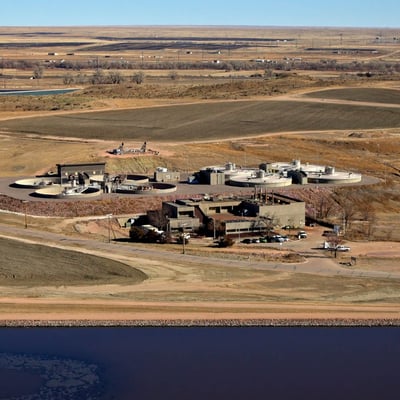 An aerial photo of a tan brick facility with circular structures emerging from a hill behind the main building. In the foreground, a dark blue lake is visible before a road. It's a sunny winter day.