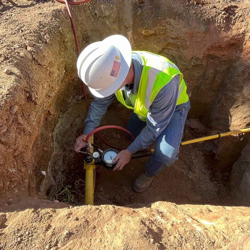 Un trabajador con equipo de seguridad en un pozo excavado utiliza un dispositivo de medición junto a una tubería amarilla de gas natural.