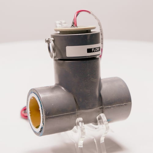 A grey T-shaped pipe connection with a lid on the top that contains a red and black wire coming from the top of the assembly.