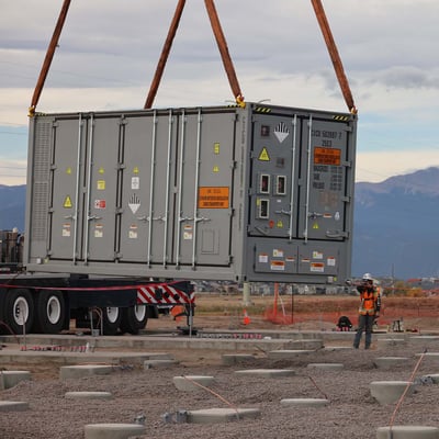 A large industrial size battery is suspended in mid-air by a crane. The battery resembled a shipping container and is painted grey. Workers in safety gear stand nearby and help guide it to the gravel lot where it is housed.