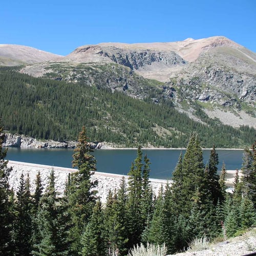 A dam lined with rock holds back water in a mountain reservoir. Tall, rugged mountains rise in the background with trees along the shoreline.