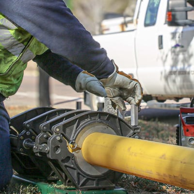 Close up photo of a worker in safety gear cutting a yellow plastic natural gas pipe. A Colorado Springs Utilities vehicle is visible in the background.