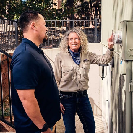 A service technician in Colorado Springs Utilities gear points out a detail on an electric meter outside of a customer's home.
