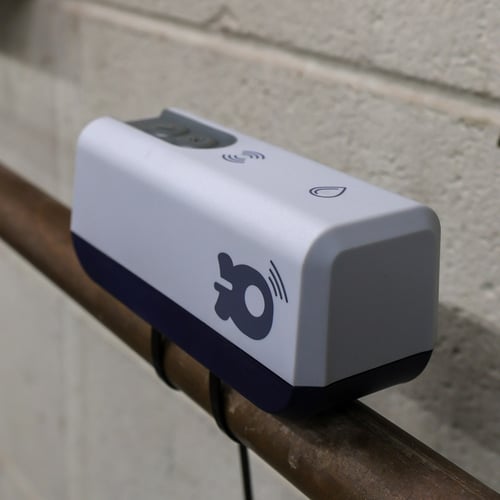 Close up shot of a grey plastic rectangle submeter strapped to a copper water pipe in an industrial room. The device has a water drop on it and a WiFi signal, indicating its connectivity as a water submeter.