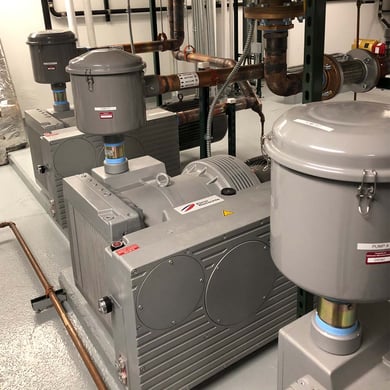 Photo of a grey oil-cooled vacuum pump system in an industrial room. Copper pipes run to and from the machinery.