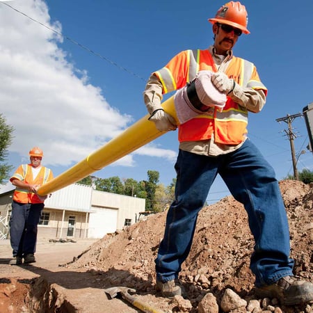 Two workers wearing safety gear carry a yellow PVC natural gas pipeline near an open trench in a street.