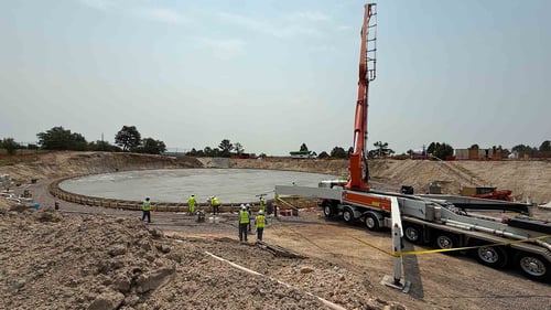 A group of workers stand next to a freshly poured flat, circular cement structure. A concrete pumper with an attached crane is parked next to them.