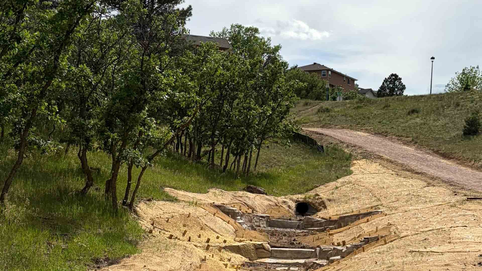 Restored drainage channel with stepped rock structures and a culvert inlet near a trail, surrounded by grass, trees and homes.