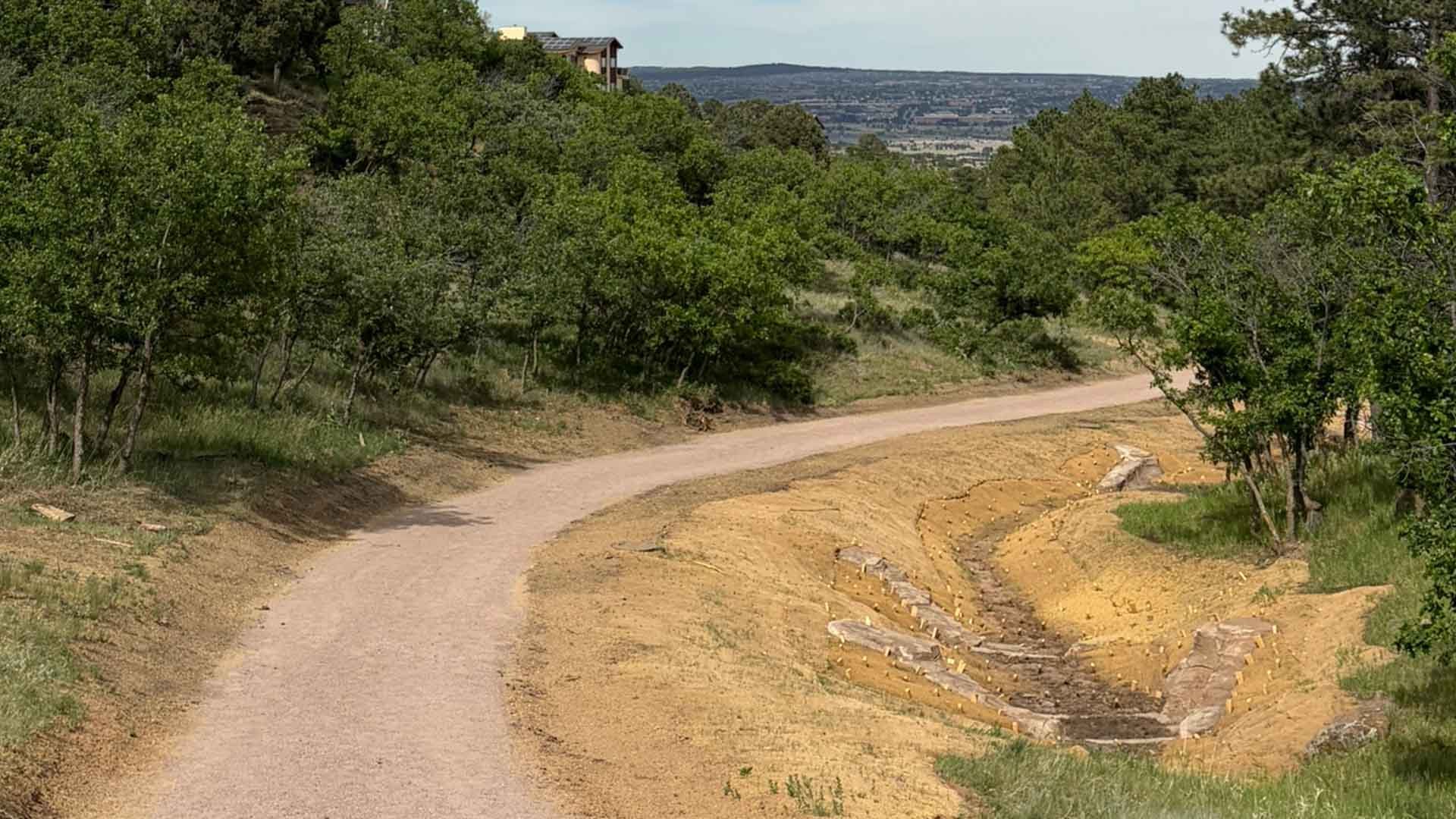 Curving trail beside a stabilized drainage channel lined with rock and erosion-control matting, surrounded by trees and open space.