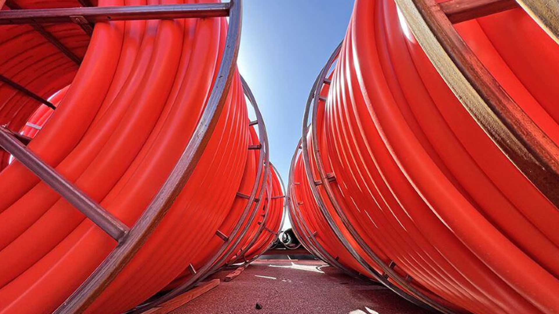 Six bright orange fiber conduit rolls stand side by side next to each other outdoors under blue sky.