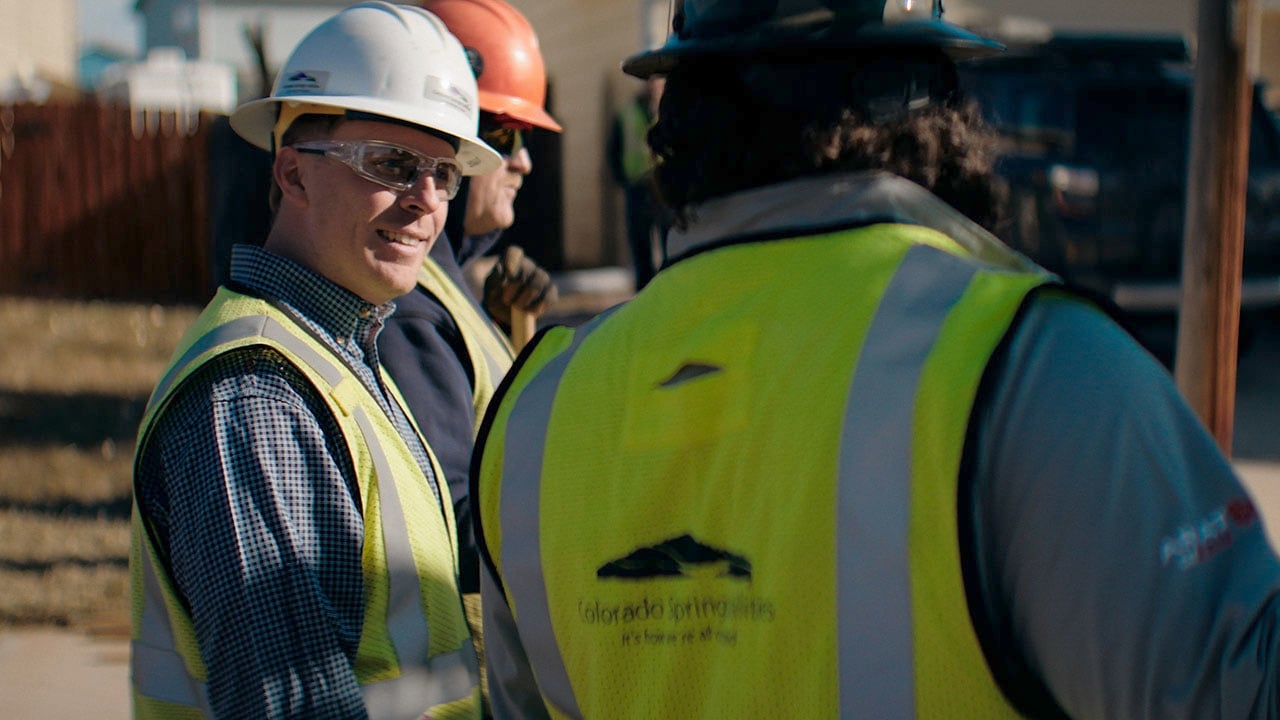 A group of three Colorado Springs Utilities natural gas employees stand talking to each other at a construction site outdoors on a residential street.