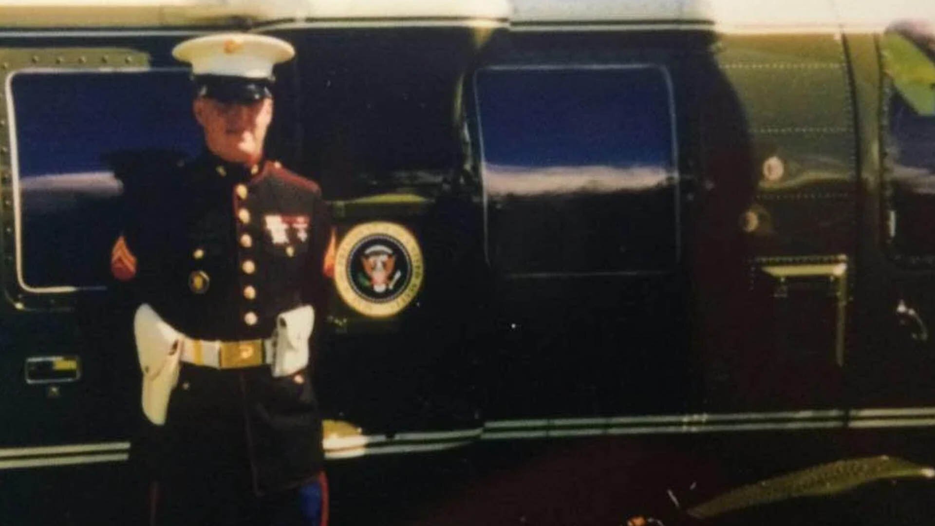 A uniformed Marine, Travas Deal, stands in front of a helicopter with the Presidential Seal on its side.