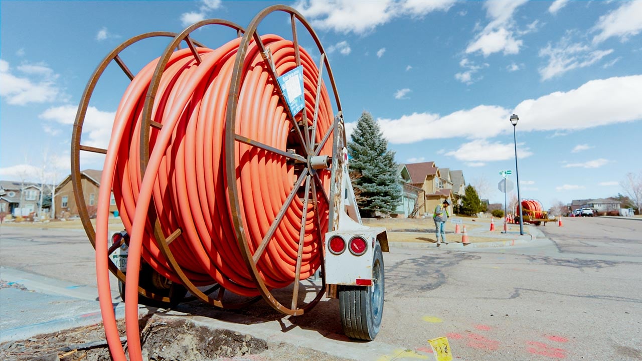 A large roll of orange fiber optic cables are mounted on a trailer parked in a residential neighborhood. A worker in safety gear is visible in the background next to orange construction cones.