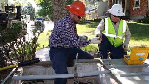 One worker, in a red hard hat and checkered shirt, is inside an underground utility box. The other kneels beside it. They are handling tools from a yellow case labeled "Multimeter."