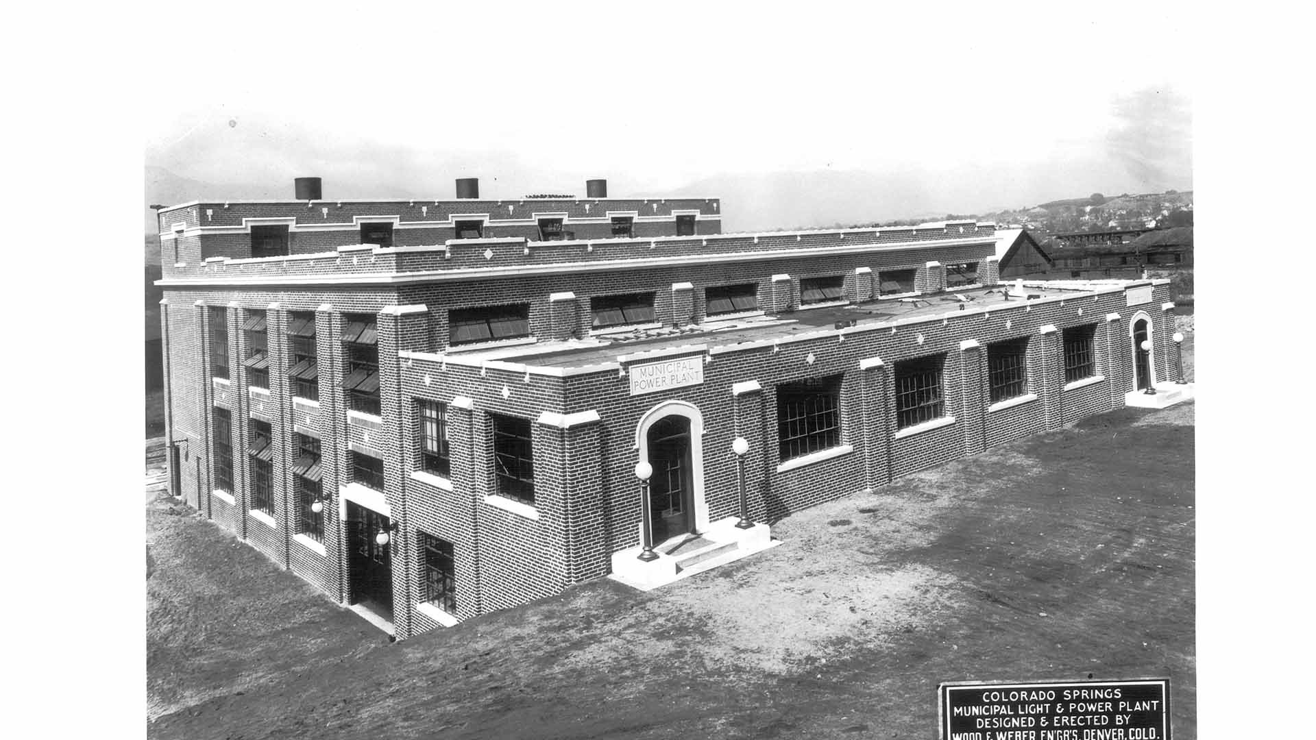 A black and white historic photo of the Martin Drake Power Plant from its early days. The plant has a brick facade and a sign above the door reads "municipal power plant"