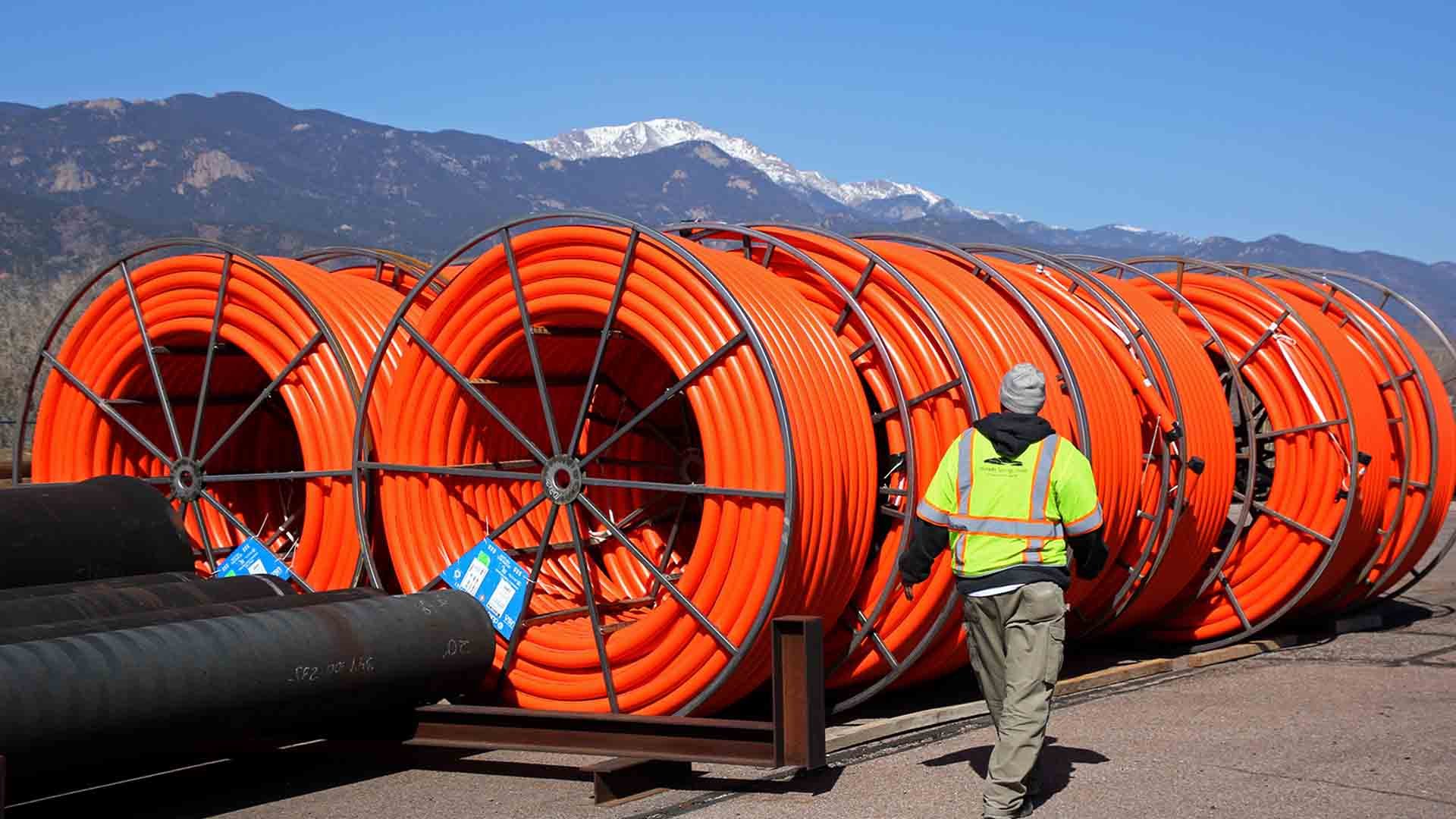 A worker in a high-visibility neon green vest walks in front of five rolls of orange fiber conduit. Pikes Peak and the front range are visible in the background.
