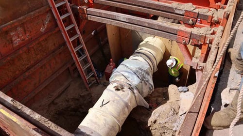 A worker in safety gear inspecting a large pipe in a hole surrounded by red metal shoring. A ladder extending into the hole is also visible.