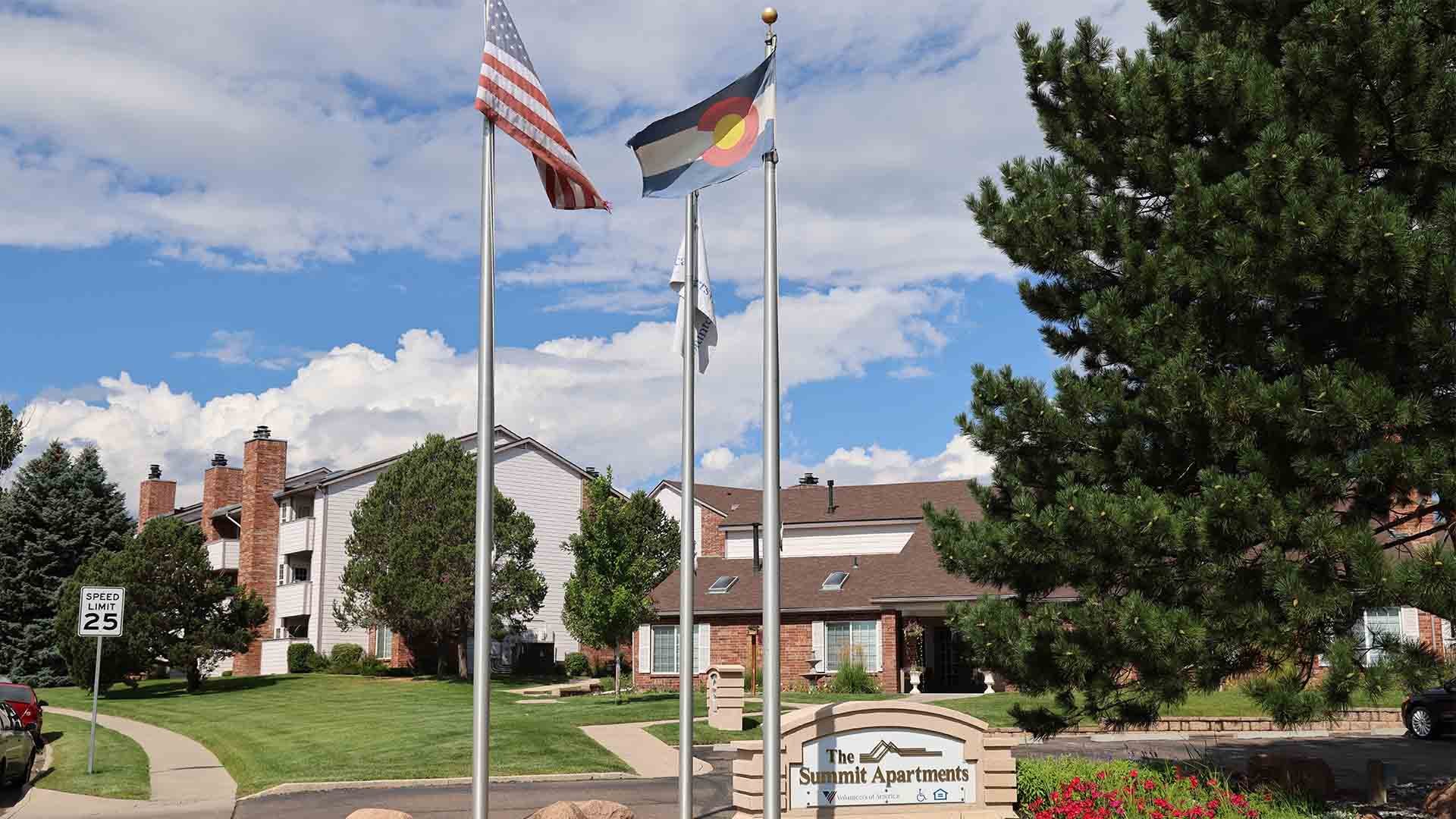 American and Colorado flags fly outside of an apartment complex. A sign in the foreground reads "The Summit Apartments."