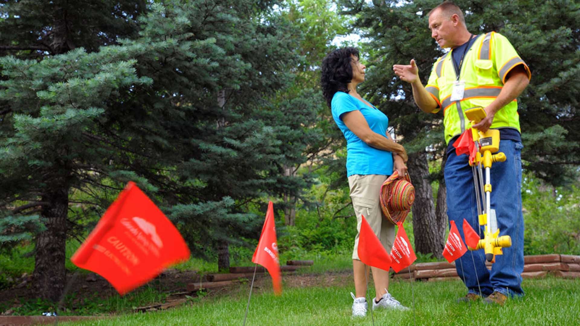 A utility worker in a high-visibility yellow vest and blue jeans stands in a grassy yard, gesturing as he speaks to a woman in a blue shirt. Several red "Call Before You Dig" flags are planted in the lawn in the foreground. The background features lush evergreen trees.