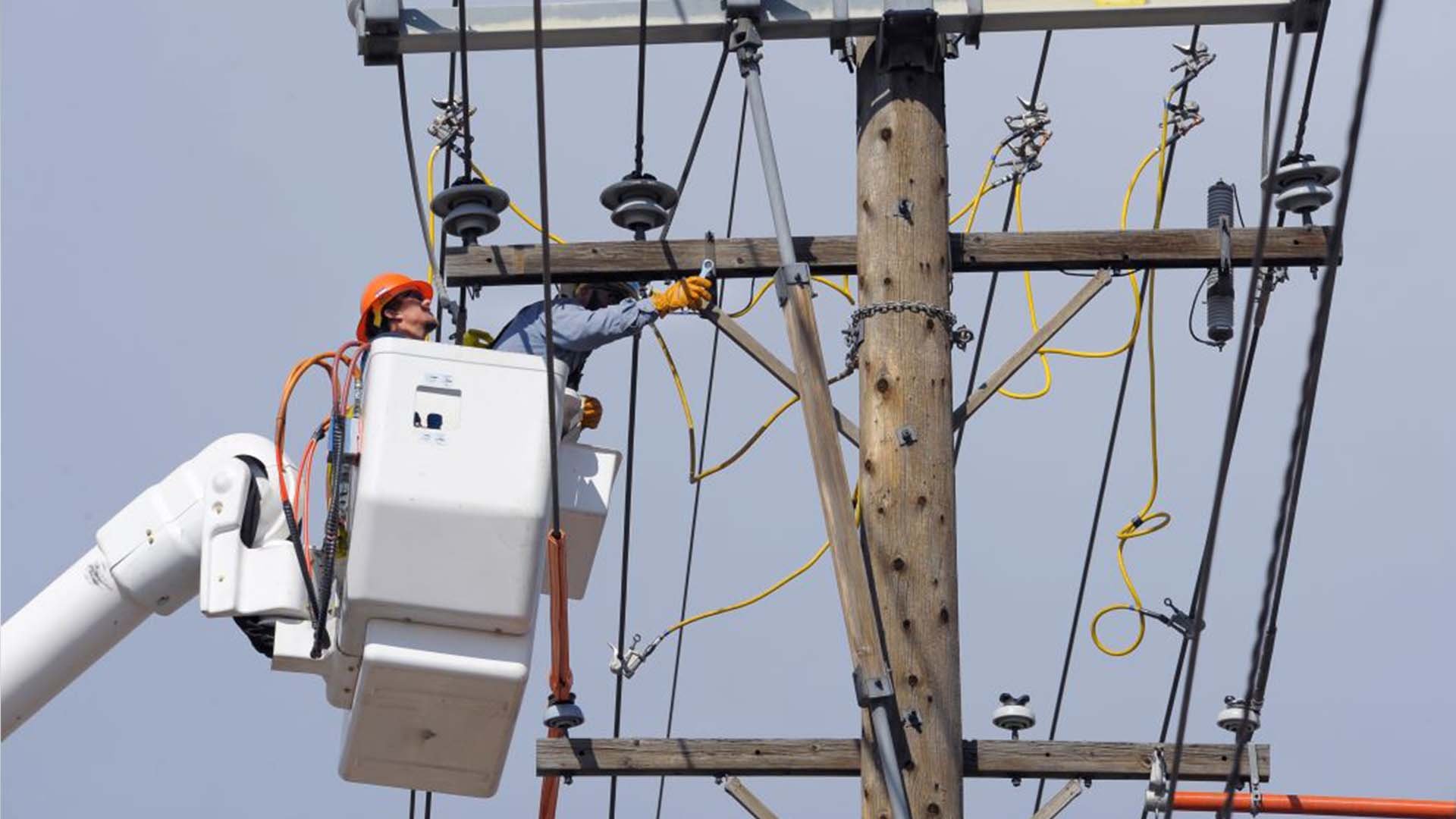 Photo of two utility workers in a bucket high above the ground maintaining electrical power lines on a wooden utility pole.