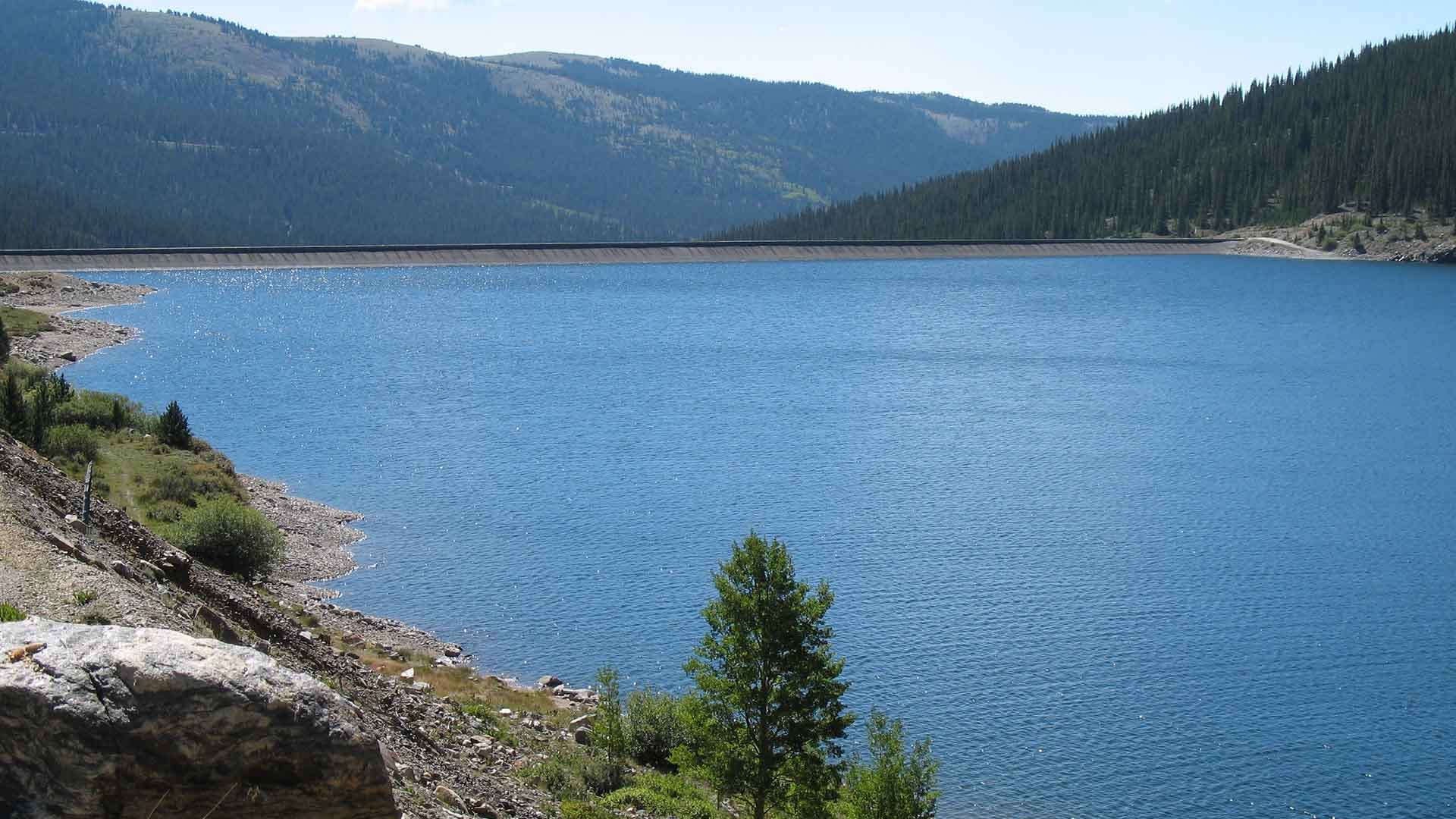 Blue waters of Montgomery Reservoir in summit county. Hills covered in trees are visible behind the reservoir's dam.