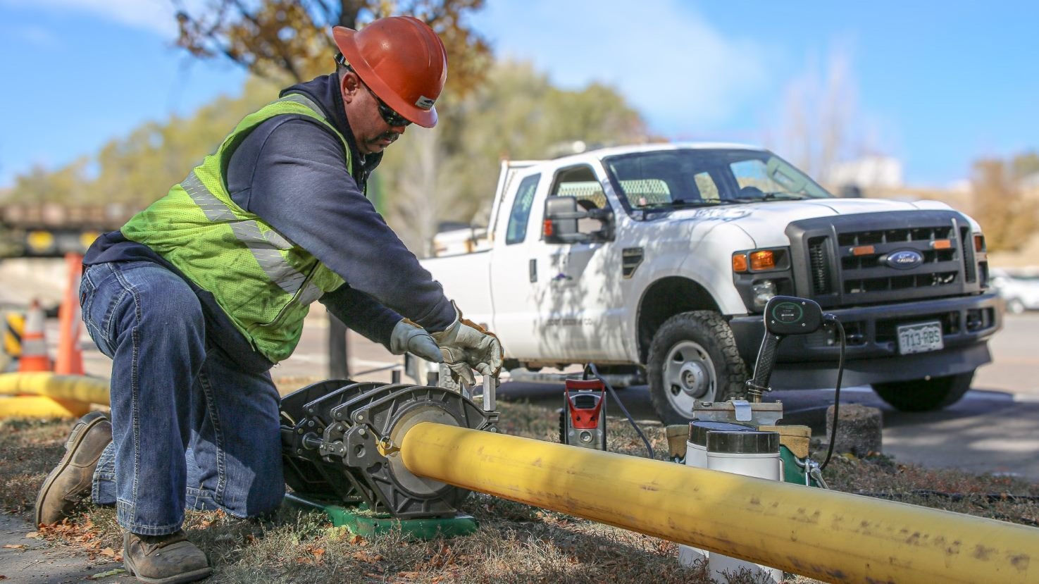 A utility worker tightening large metal clamps onto a yellow gas line. A white truck is in the background.