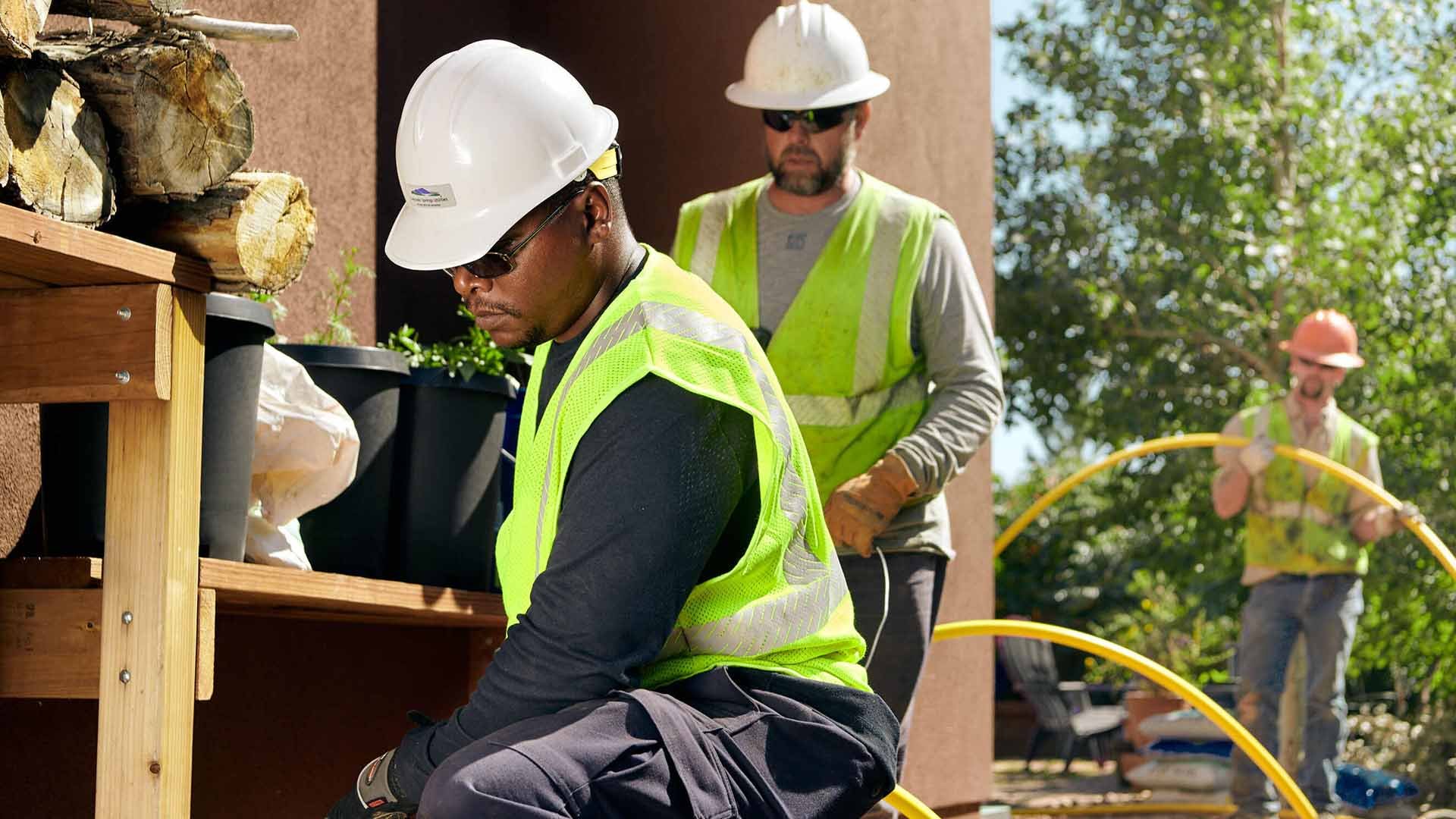 Two workers in safety vests and hard hats handle a flexible yellow piece of conduit outdoors next to a home.