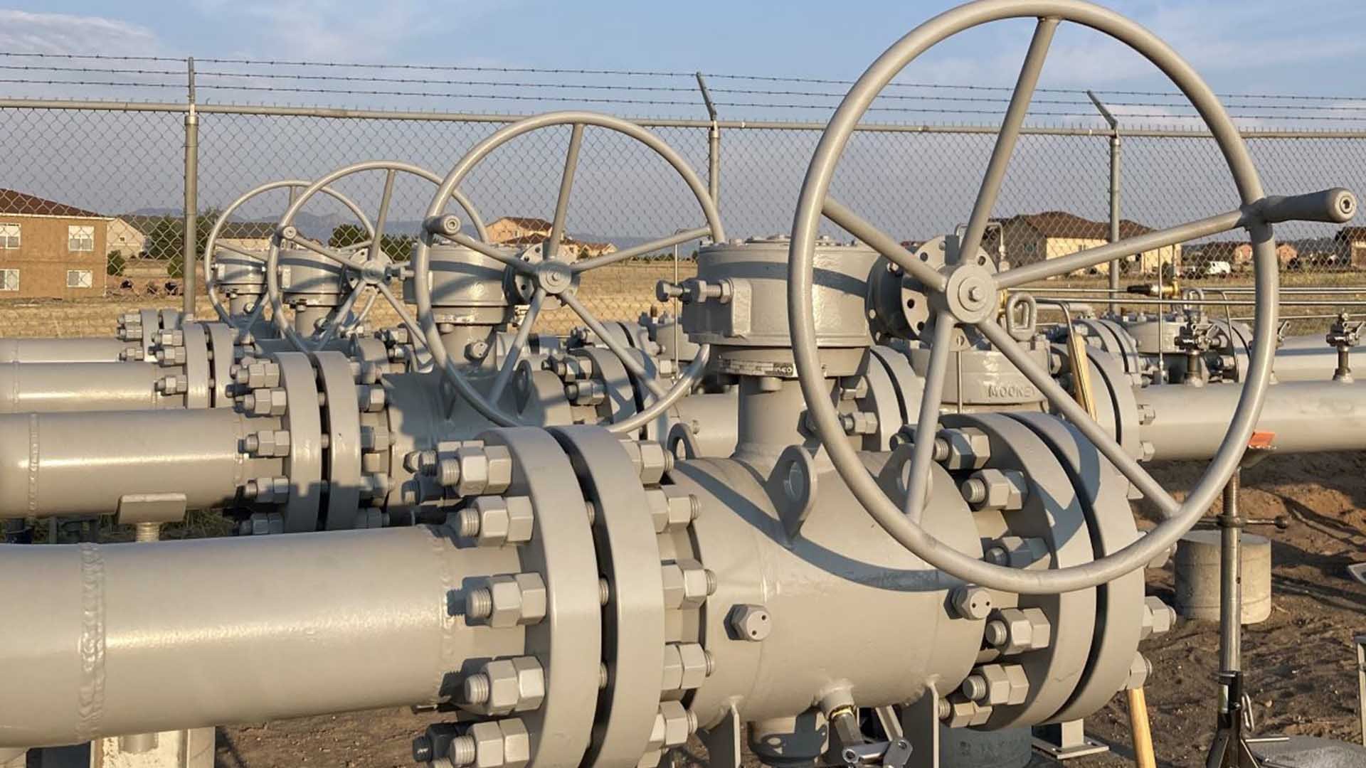 Four large wheels that control the flow of natural gas attached to grey pipelines photographed in the morning sun.