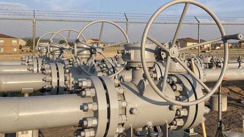 Four large wheels that control the flow of natural gas attached to grey pipelines photographed in the morning sun.