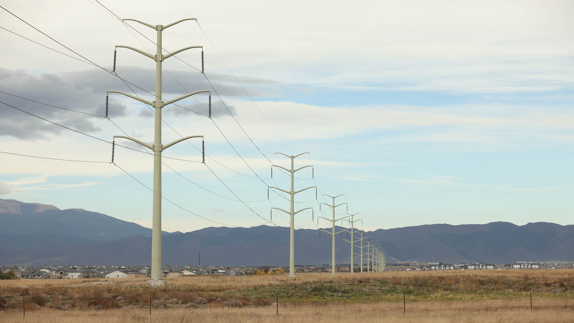 A series of large transmission lines stretching across a dry, grassy field with mountains in the background under a partly cloudy sky.