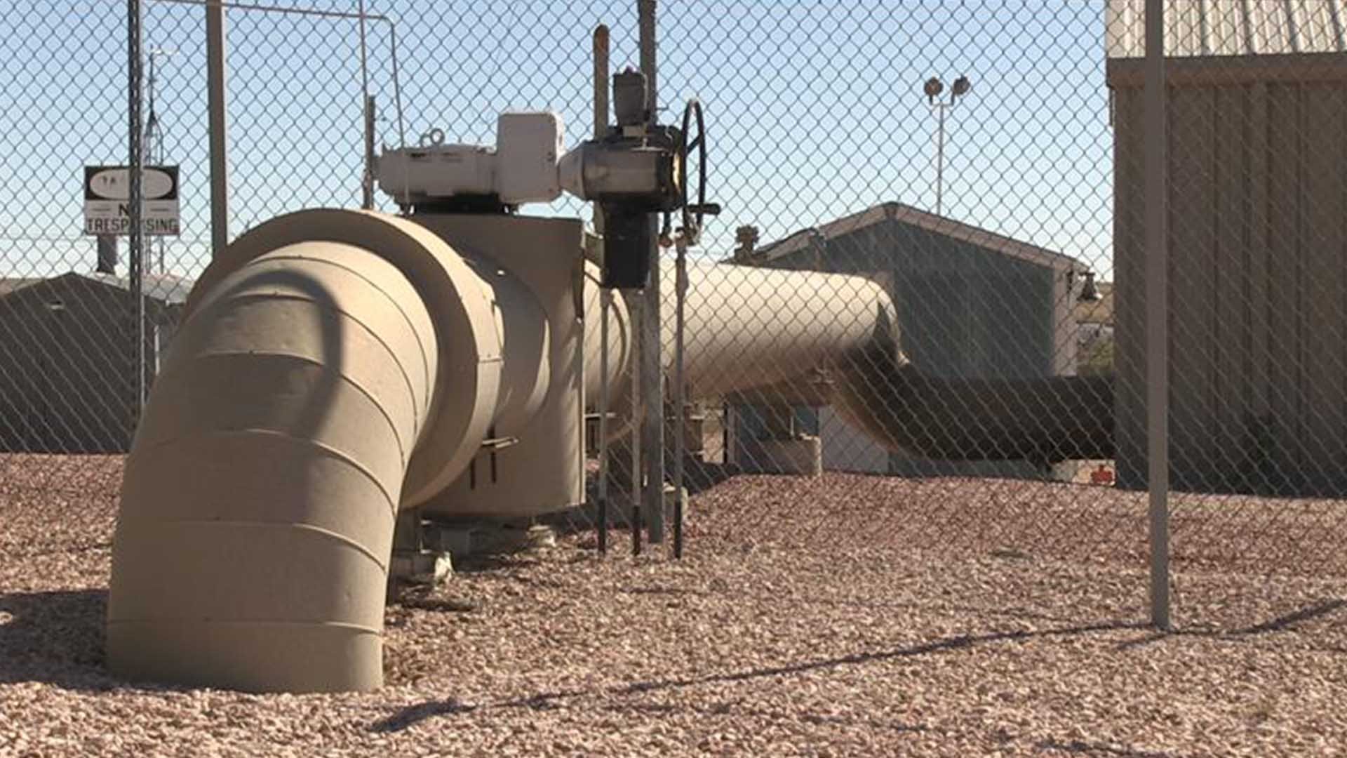 A very large tan natural gas pipe section that runs from the ground to a building behind a fence.