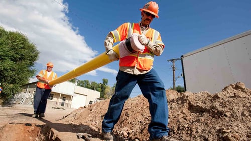Two workers wearing safety gear carry a yellow PVC natural gas pipeline near an open trench in a street.