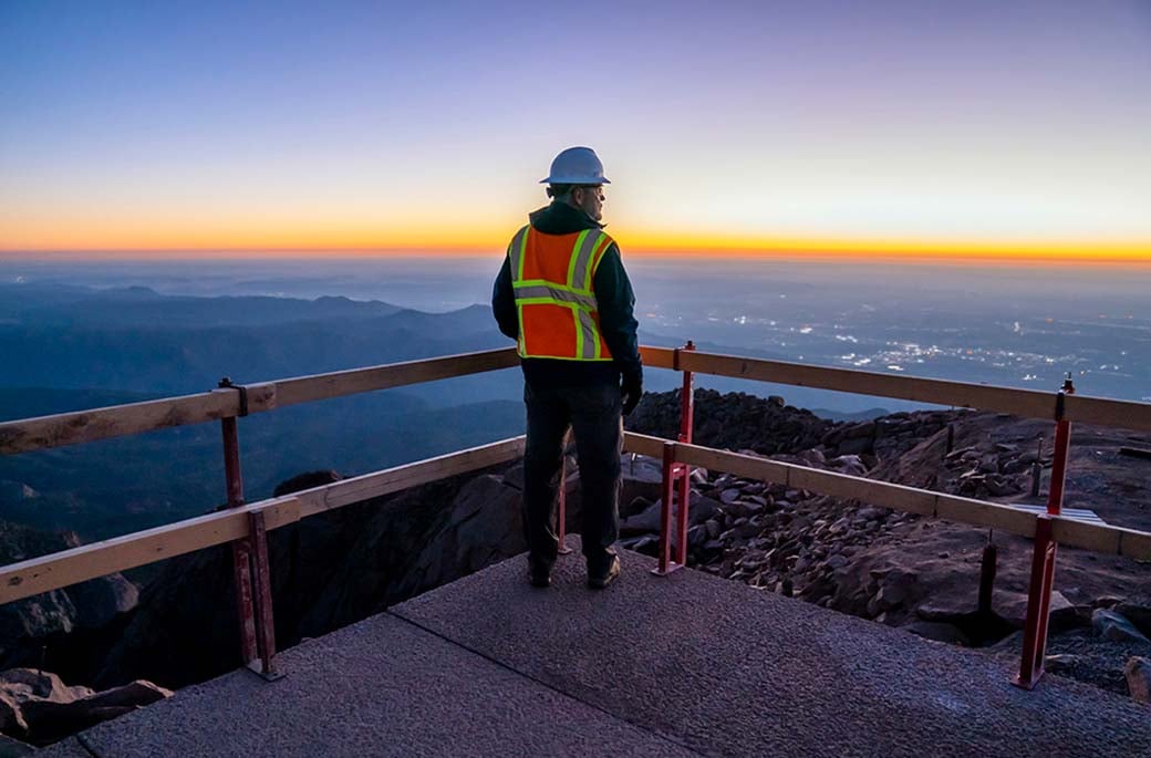 Someone wearing an orange, high-visibility, vest facing the sunset while standing on a platform on a mountain peak. 