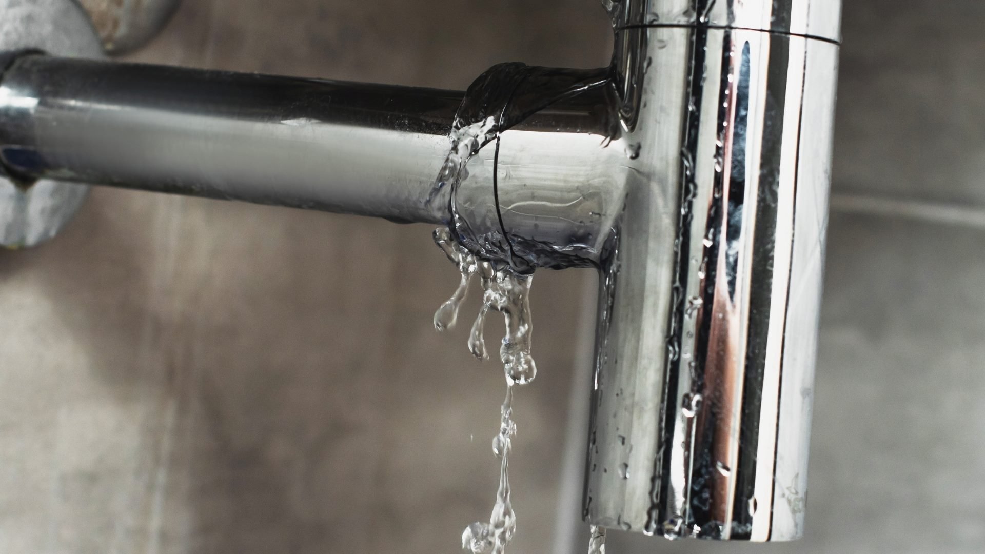 Silver pipe under a sink leaking water at the joint. The room has cement walls.