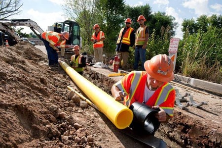 Six workers wearing safety gear stand around a trench in a street for a natural gas line.