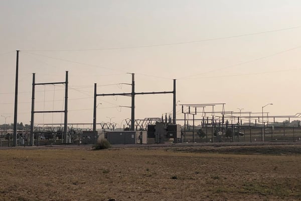 A photograph of a substation in the midle of a field. There are transmission lines running overhead. 