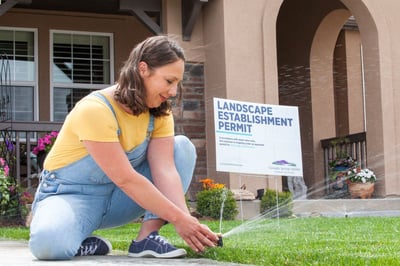 A person kneels down next to a sprinkler head spraying water on a lawn. There is a sign that reads "Landscape Establishment Permit" in the yard.