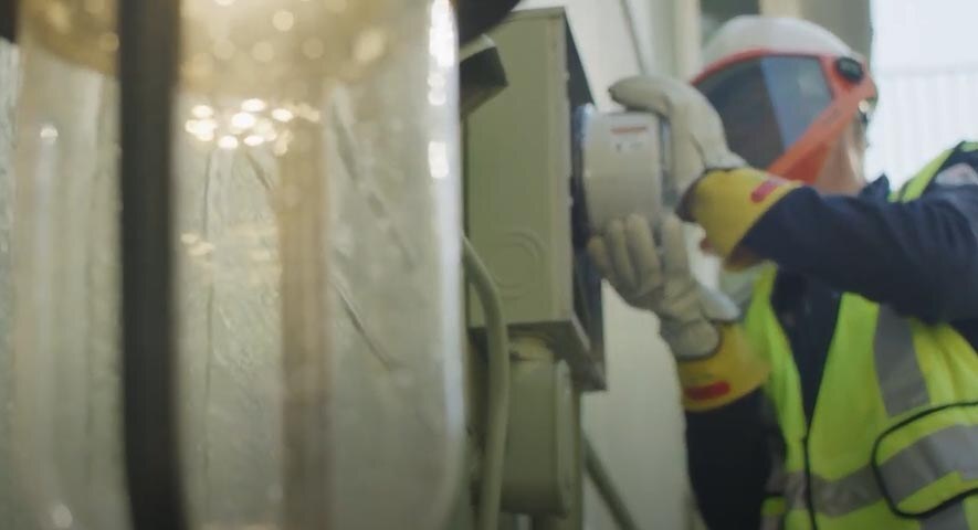 A person in a high-visibility vest and protective helmet with a face shield is replacing the utilities meter on the side of a home.
