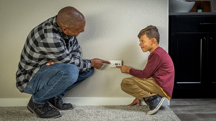 A father and son are squatting low against a light wall near a carpeted floor. The father, a Black man, is wearing a plaid shirt and pointing to a carbon monoxide detector. The son, a young boy, is wearing a long-sleeve shirt and is also pointing at the controls with a curious expression.