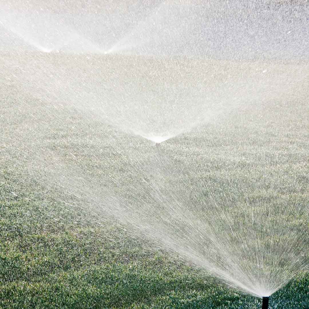 Four in-ground sprinklers spraying green grass with water in a cone pattern.